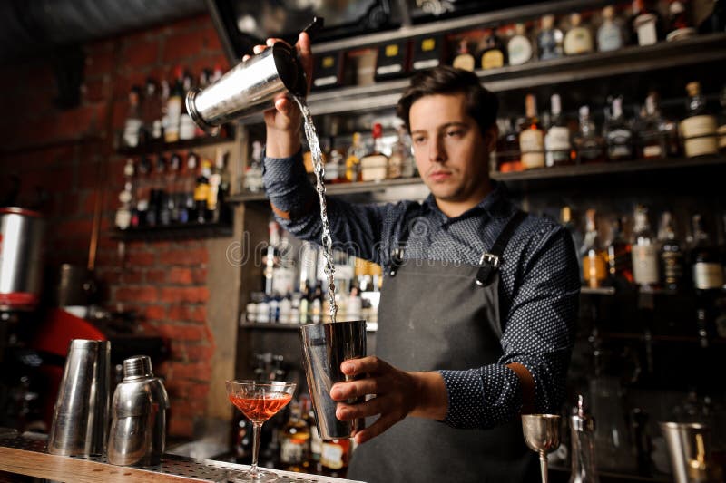 Junger Kellner, der alkoholisches Getränk von einem Metallglas in andere gießt lizenzfreies stockbild Junger Kellner, der alkoholisches Getränk von einem Metallglas in andere gießt lizenzfreies stockbild
