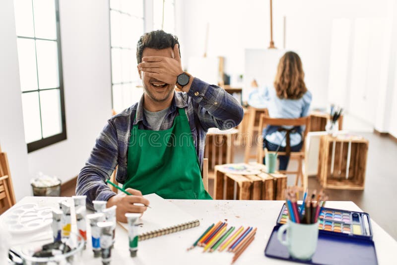 Young artist man at art studio smiling and laughing with hand on face covering eyes for surprise. blind concept. Young artist man at art studio smiling and laughing with hand on face covering eyes for surprise. blind concept