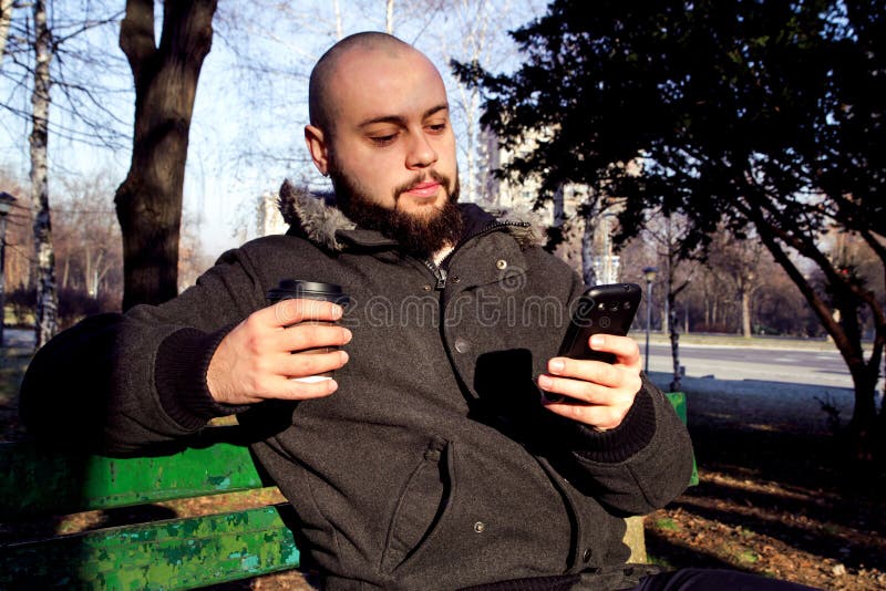 Junger Mann, der im Stadtpark mit intelligentem Telefon und dem Trinken sitzt lizenzfreie stockfotos Junger Mann, der im Stadtpark mit intelligentem Telefon und dem Trinken sitzt lizenzfreie stockfotos