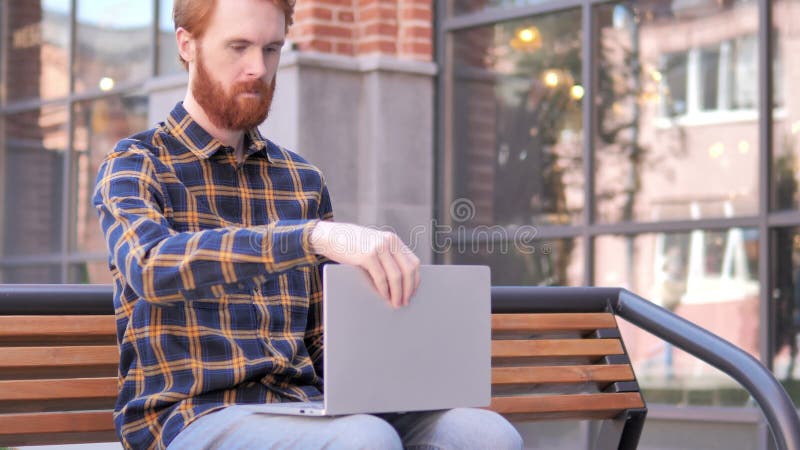 Junger Mann des Rothaarigebartes, der auf Bank kommt und sitzt, Laptop zu benutzen