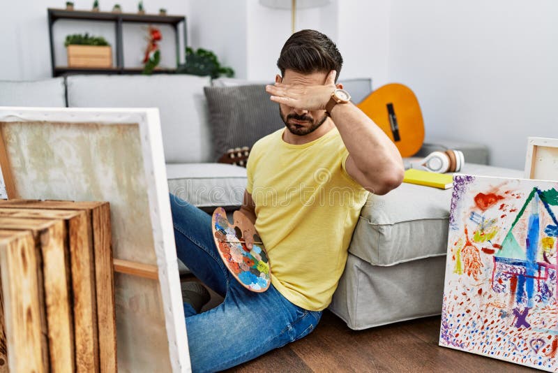 Young man with beard painting canvas at home covering eyes with hand, looking serious and sad. sightless, hiding and rejection concept. Young man with beard painting canvas at home covering eyes with hand, looking serious and sad. sightless, hiding and rejection concept