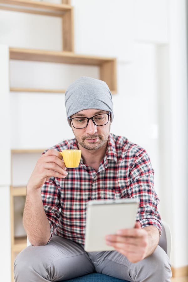 Junger modischer Mann, der im Büro sitzt und seinen Morgenkaffee beim Ablesen von Nachrichten auf Tablettengerät genießt lizenzfreies stockbild Junger modischer Mann, der im Büro sitzt und seinen Morgenkaffee beim Ablesen von Nachrichten auf Tablettengerät genießt lizenzfreies stockbild