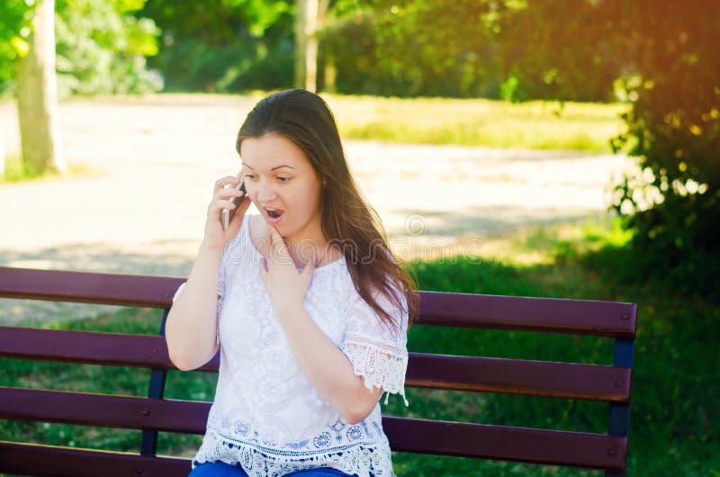 Junger schöner europäischer Mädchen Brunette sitzend auf einer Bank in einem Stadtpark und sprechend am Telefon und überrascht gu lizenzfreie stockfotos Junger schöner europäischer Mädchen Brunette sitzend auf einer Bank in einem Stadtpark und sprechend am Telefon und überrascht gu lizenzfreie stockfotos