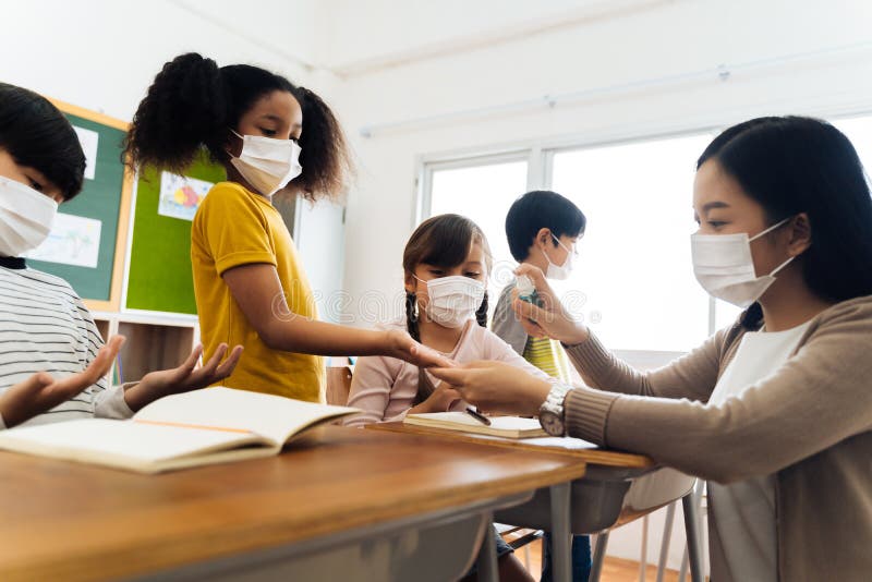 Junger weiblicher Lehrer mithilfe eines Alkoholsprays, Studentenhänden im Klassenzimmer zu desinfizierendem in der Gesichtsmasker