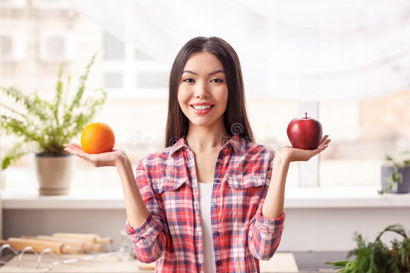 Junges Mädchen an der gesunden Lebensstilstellung der Küche, die den Apfel und Orange das frohe Vergleichen der Kamera schauend h lizenzfreies stockfoto Junges Mädchen an der gesunden Lebensstilstellung der Küche, die den Apfel und Orange das frohe Vergleichen der Kamera schauend h lizenzfreies stockfoto