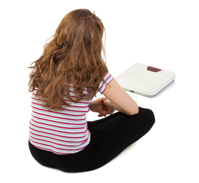 Young girl sitting sadly on the floor and looking at the scale in front of her, isolated on white. Young girl sitting sadly on the floor and looking at the scale in front of her, isolated on white.