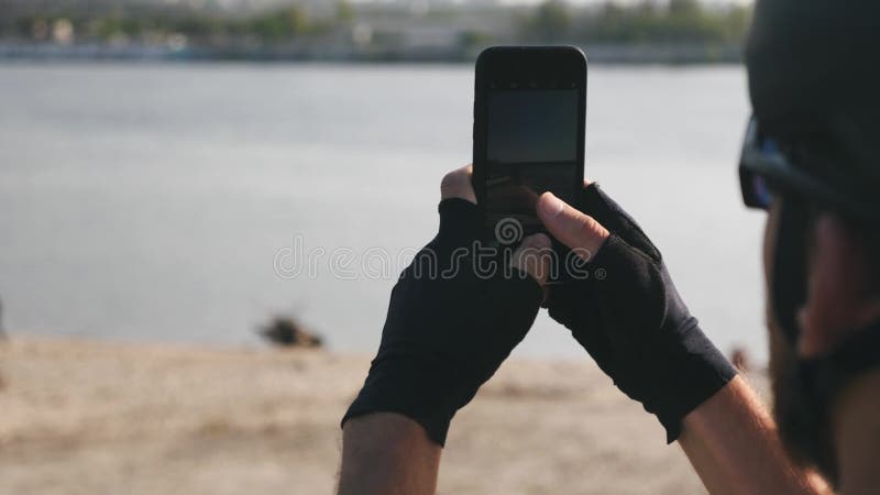 Junges Radfahrerholdingtelefon in seiner Hand, die Fotos des tragenden Sturzhelms des Flusses und der Stadt, der Sonnenbrille und