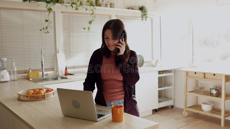 Jóven independiente trabajando en laptop mientras habla por teléfono móvil desde la cocina almacen de video