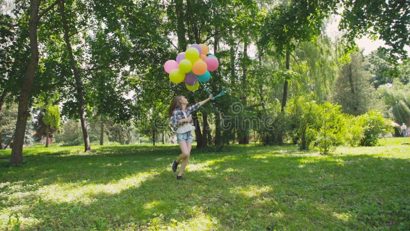 Júbilo feliz de la chica joven y giro con los globos coloridos en un parque almacen de metraje de vídeo