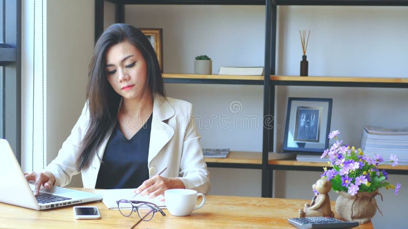 4K lengte, bezige bedrijfsvrouw die met laptop computer werken, op boek schrijven en met ernstig gezicht bij het werkruimte denke