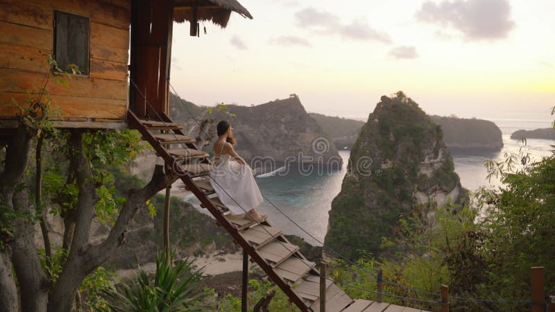 4k видео женщины белое платье сидеть дома дерева nusa penida bali indonesia. акции видеоматериалы