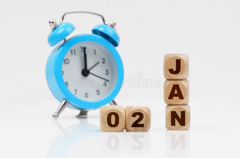 2st of January. Wooden cube calendar for January 2, next to a blue alarm clock. Objects on a reflective white surface. 2st of January. Wooden cube calendar for January 2, next to a blue alarm clock. Objects on a reflective white surface