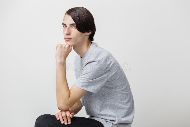 Kalme en ontspannen jonge dunne donker-haired kerel met blauwe ogen die grijze t-shirt dragen, die op stoel tegen wit zitten royalty-vrije stock afbeeldingen Kalme en ontspannen jonge dunne donker-haired kerel met blauwe ogen die grijze t-shirt dragen, die op stoel tegen wit zitten royalty-vrije stock afbeeldingen