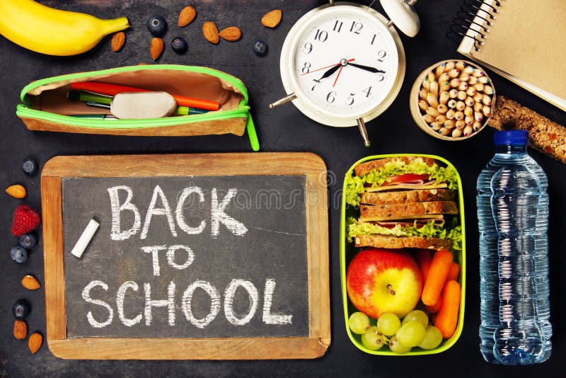 Sandwich, apple, grape, carrot, berry in plastic lunch boxes, stationery and bottle of water on black chalkboard. Back to school concept. Sandwich, apple, grape, carrot, berry in plastic lunch boxes, stationery and bottle of water on black chalkboard. Back to school concept.