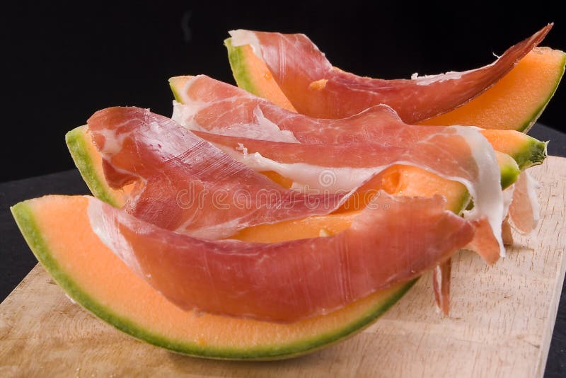 Sliced cantaloupe draped with prosciutto on wooden board, black background. Sliced cantaloupe draped with prosciutto on wooden board, black background.