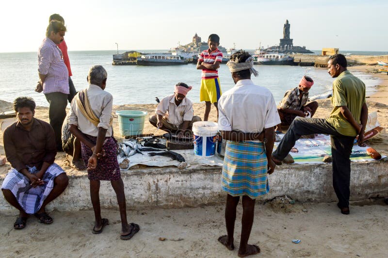 Kanyakumari, Tamil Nadu, India. 08/14/14. A man selling fish on the edge of the beach as people look on, with the statue of Viveka stock photos Kanyakumari, Tamil Nadu, India. 08/14/14. A man selling fish on the edge of the beach as people look on, with the statue of Viveka stock photos