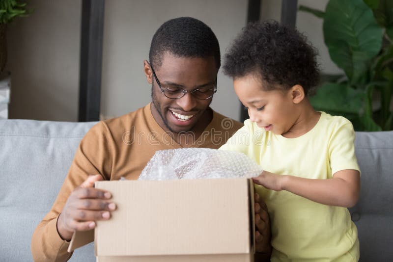 Happy black father and little kid son opening cardboard box looking inside, african american dad with cute child toddler clients receiving carton package, post mail parcel delivery service concept. Happy black father and little kid son opening cardboard box looking inside, african american dad with cute child toddler clients receiving carton package, post mail parcel delivery service concept