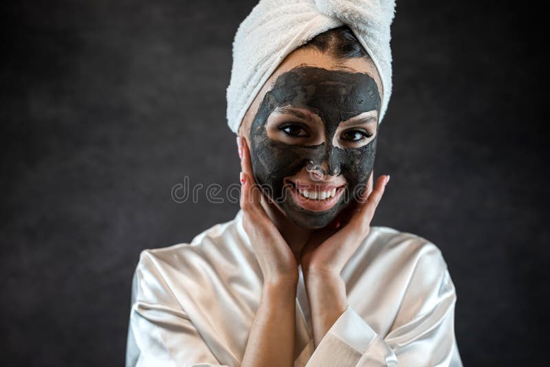 Kaukasierin trägt weißes Handtuch mit schwarzer Lehmmaske, die auf schwarzem Lehm isoliert ist lizenzfreie stockfotografie