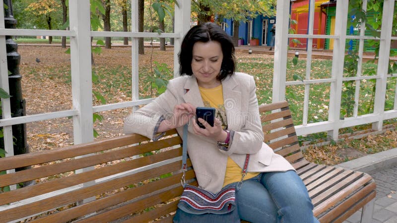 Kaukasische brunette vrouw van middelbare leeftijd die op een parkbank zit met telefoon