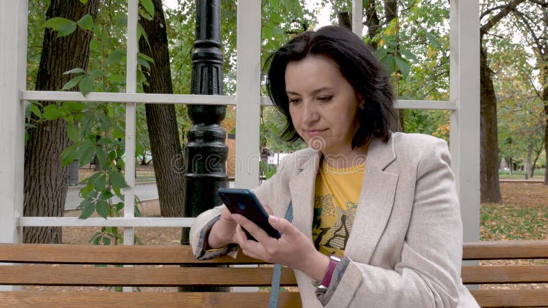 Kaukasische brunette vrouw van middelbare leeftijd die op een parkbank zit met telefoon