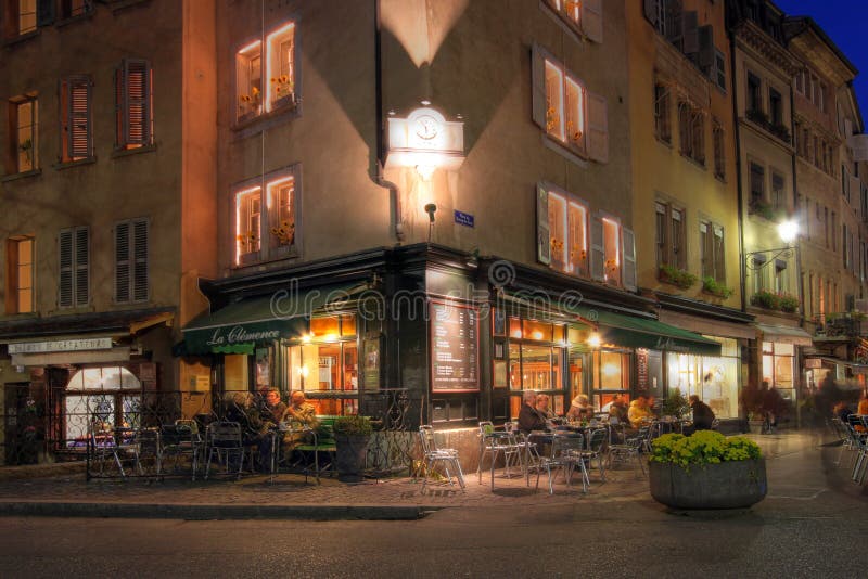 Place de Bourg is the heart of the old town in Geneva, Switzerland. The numerous terraces offering drinks and food are very popular for tourists and locals alike. This night-time photograph (HDR) is centered on a corner coffee-shop. The image was taken during late autumn, hence people are wearing thick clothing. Place de Bourg is the heart of the old town in Geneva, Switzerland. The numerous terraces offering drinks and food are very popular for tourists and locals alike. This night-time photograph (HDR) is centered on a corner coffee-shop. The image was taken during late autumn, hence people are wearing thick clothing.