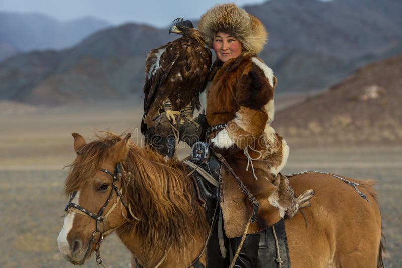 SAGSAY, MONGOLIA - SEP 28, 2017: Kazakh woman Eagle Hunter traditional clothing, while hunting to the hare holding a golden eagle on his arm in desert mountain of Western Mongolia. SAGSAY, MONGOLIA - SEP 28, 2017: Kazakh woman Eagle Hunter traditional clothing, while hunting to the hare holding a golden eagle on his arm in desert mountain of Western Mongolia.