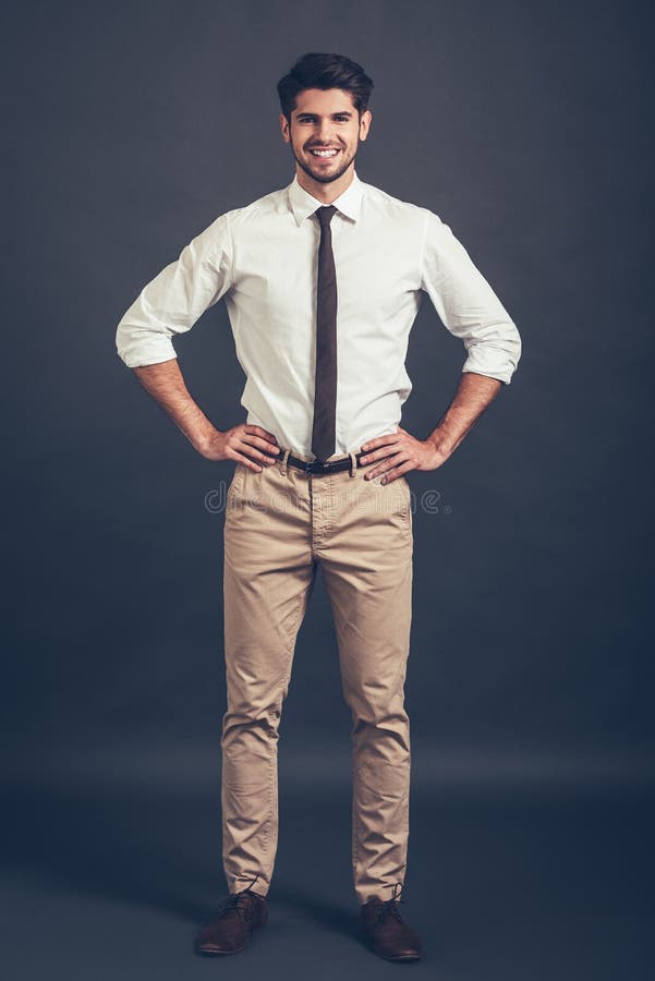 Full length of confident young handsome man keeping arms akimbo and looking at camera with smile while standing against grey background. Full length of confident young handsome man keeping arms akimbo and looking at camera with smile while standing against grey background