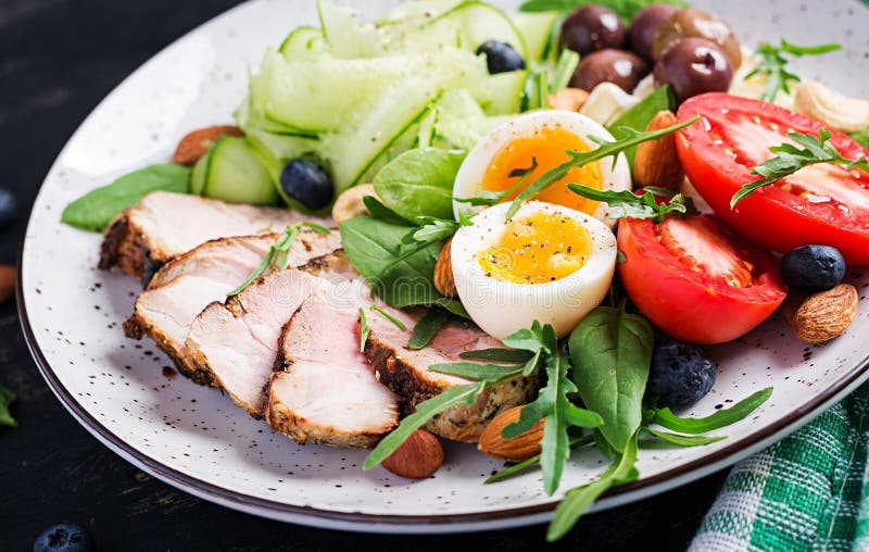 Ketogenic diet. Boiled egg, pork steak and olives, cucumber, spinach, brie cheese, nuts and tomato