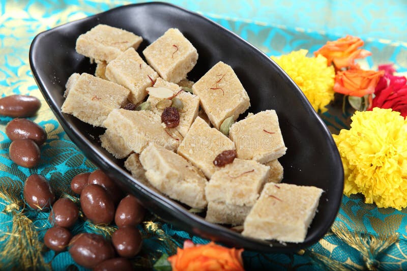 In this image a black bowl overflows with delectable Khoya Burfi, a traditional Indian sweet. The bowl is placed on a vibrant green patterned background, and surrounding, along with colorful candies and a delicate rose flowers as a beautiful garnish. Khoya Burfi, known by various names such as Mathura Peda, Mawa Barfi, Kowa Barfi, and more, is a beloved dessert originating from Mathura, Uttar Pradesh in India.To create this mouthwatering treat, the main ingredients used are khoa (also known as khoya) and sugar. Khoya is a dairy product made by reducing milk until it becomes a solid mass. The recipe for Khoya Burfi is delightfully simple and quick, making it a perfect choice for parties or gifting.Let's dive into the ingredients needed for Easy Khoya Burfi: 1 cup of grated Khoya or Mawa, 3 tablespoons of sugar, 1 teaspoon of ghee (clarified butter), � teaspoon of cardamom powder (also known as Yelakai podi), 3 tablespoons of assorted nuts (such as pista, badam, and cashews), a pinch of saffron, and 1 tablespoon of milk.Now, let's move on to the preparation process. Begin by lining a plate with foil and greasing it generously with ghee. Set the prepared plate aside. In a pan, heat the ghee and add the assorted nuts. Fry them until they turn golden, then remove them from the pan and set them aside.Next, soak the saffron in a tablespoon of milk for about 5 minutes and set it aside. Now, add the grated khoya to the same pan and fry it for a few seconds. Add the sugar and mix well. Continue cooking the mixture until it starts to leave the sides of the pan. At this point, add the cardamom powder, saffron milk, and the fried nuts. Give everything a good mix.Pour the mixture into the greased plate and smoothen the surface. Sprinkle some additional nuts on top for added visual appeal. Allow the Khoya Burfi to set for about an hour. Once set, cut it into squares and savor each bite.For storage, you can refrigerate the Khoya Burfi and enjoy its freshness for 4 to 7 days. With its rich flavors and delightful texture, Khoya Burfi is a perfect choice for indulging in a traditional Indian sweet that's both delicious and visually appealing.