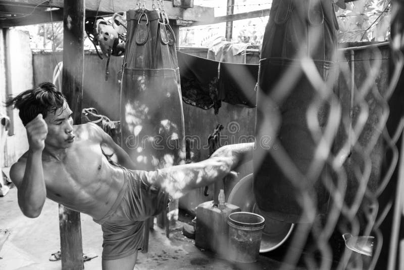 A kickboxer practising his leg kicks against a punch bag shot through a chain link fence and in black and white, Siem Reap