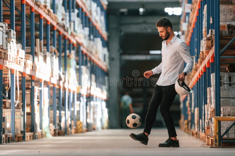 Kicking Soccer Ball. Storage Worker is in the Warehouse with Bunch of