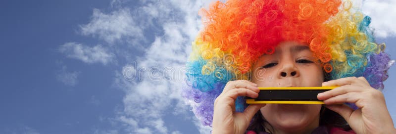 Clown kid with a colorful wig on her head, playing the harmonica under the blue clear sky. Clown kid with a colorful wig on her head, playing the harmonica under the blue clear sky.