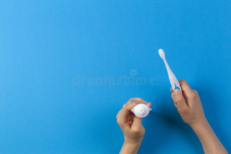 Kid hand holding electric toothbrush and putting paste from toothpaste tube over blue background