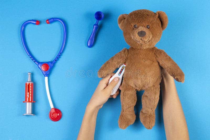 Kid hands measuring temperature with toy thermometer of teddy bear, toys medicine tools on light blue background. Top