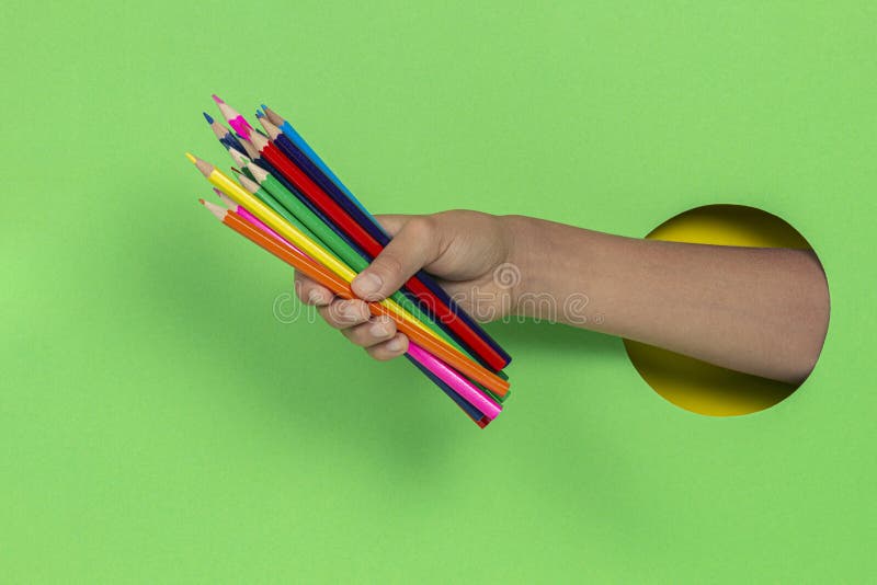 Kid holding colored pencils in hand through hole on light green background royalty free stock photos