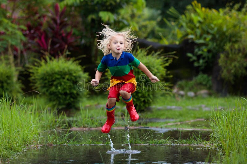 Kid playing out in the rain. Children with umbrella and rain boots play outdoors in heavy rain. Little boy jumping in muddy puddle. Kids fun by rainy autumn weather. Child running in tropical storm