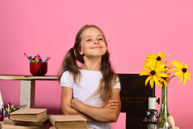 Kid and school supplies on pink wall background. Homework and back to school concept. Schoolgirl with happy proud face in classroom. Girl stands at her desk with colorful stationery, books and flowers