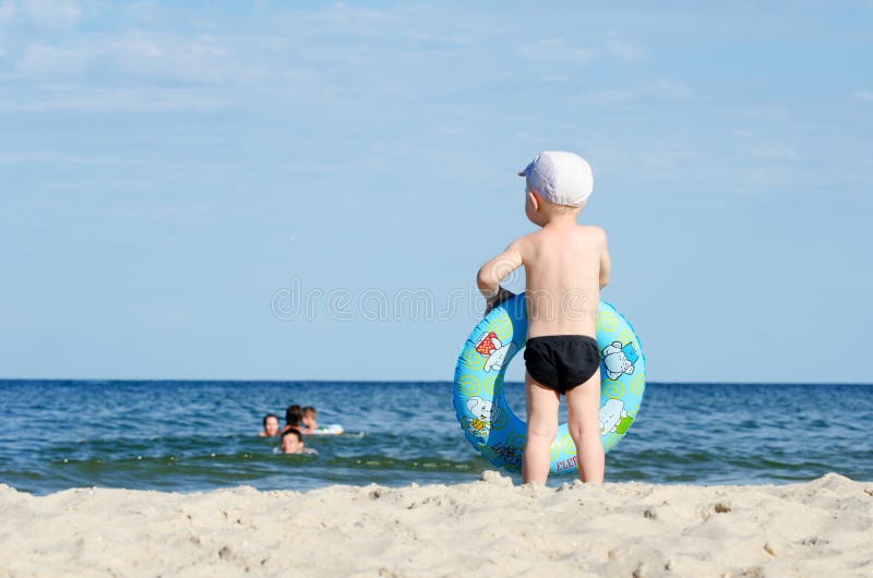 Kid in swimming trunks and a cap with a swimming circle stands on the shore of the sea back