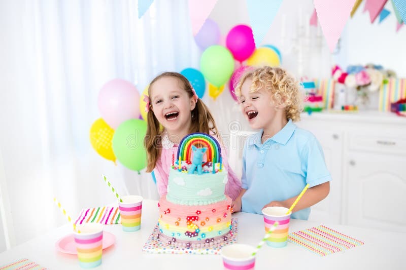 Kids birthday party with colorful pastel decoration and rainbow cake. Girl and boy with sweets, candy and fruit. Balloons and banner at festive decorated table for child or baby birthday party. Kids birthday party with colorful pastel decoration and rainbow cake. Girl and boy with sweets, candy and fruit. Balloons and banner at festive decorated table for child or baby birthday party.