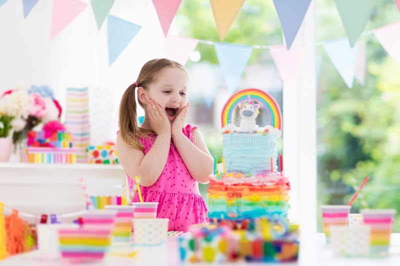 Kids birthday party with colorful pastel decoration and unicorn rainbow cake. Little girl with sweets, candy and fruit. Balloons and banner at festive decorated table for child or baby birthday party. Kids birthday party with colorful pastel decoration and unicorn rainbow cake. Little girl with sweets, candy and fruit. Balloons and banner at festive decorated table for child or baby birthday party.