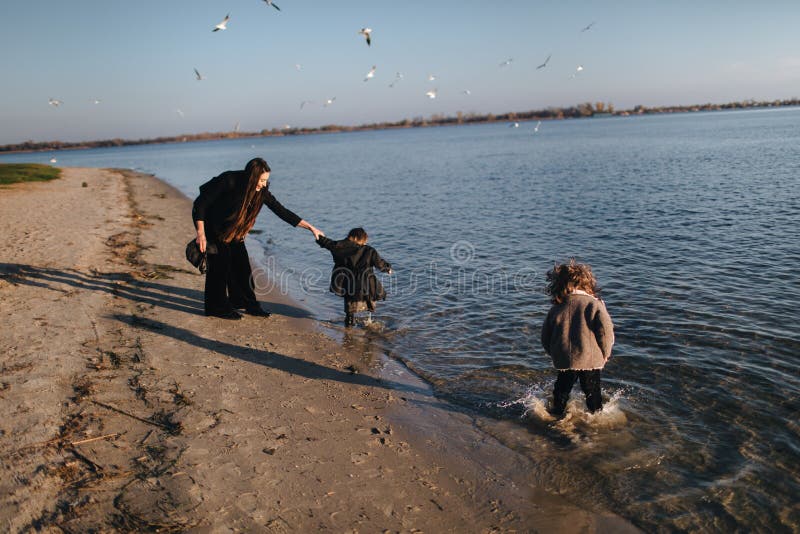 Kids and mom running in water on river bank in autumn. Boy is splashing in river water. Disobedience. Happy family on