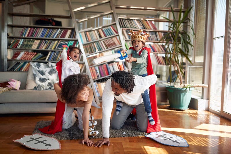 Kids are shouting while riding their parents in a knights game in a playful atmosphere at home. Family, home, love, together. Kids are shouting while riding their parents in a knights game in a playful atmosphere at home. Family, home, love, together