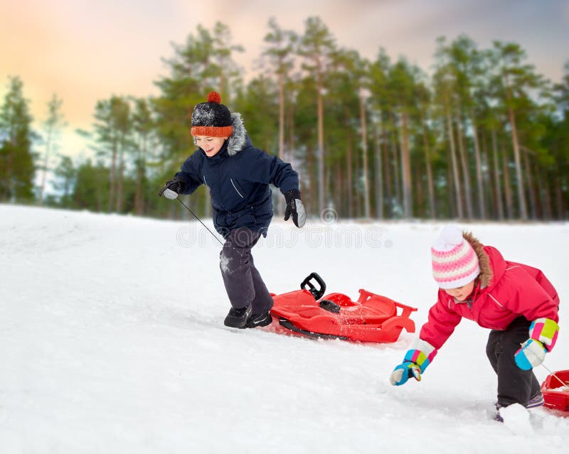 childhood, sledging and season concept - happy little kids with sleds climbing snow hill in winter over pine forest on background