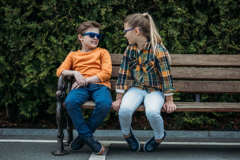 Funny kids in sunglasses talking while sitting on bench at park