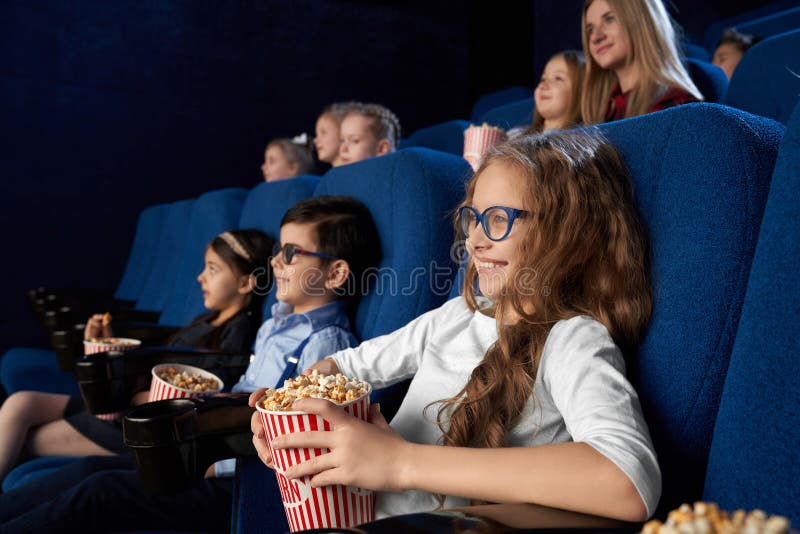 Happy, joyful kids sitting in dark blue, comfortable chairs in modern cinema theatre, watching movie or cartoon. Children enjoying film, smiling, holding buckets with popcorn. Happy, joyful kids sitting in dark blue, comfortable chairs in modern cinema theatre, watching movie or cartoon. Children enjoying film, smiling, holding buckets with popcorn.