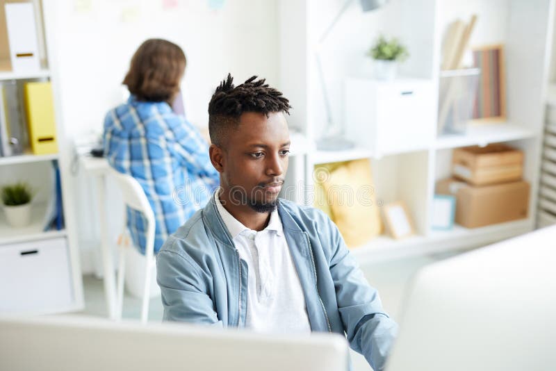 Young African-american men sitting by workplace in front of two desktop computers wotking with database. Young African-american men sitting by workplace in front of two desktop computers wotking with database