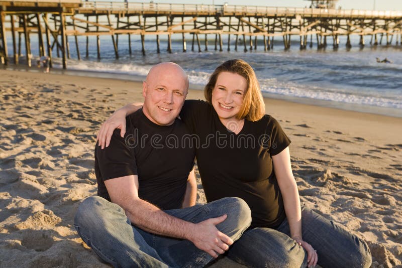 Young Pregnant Couple on the Beach with Pier in Background at Sunset. Young Pregnant Couple on the Beach with Pier in Background at Sunset