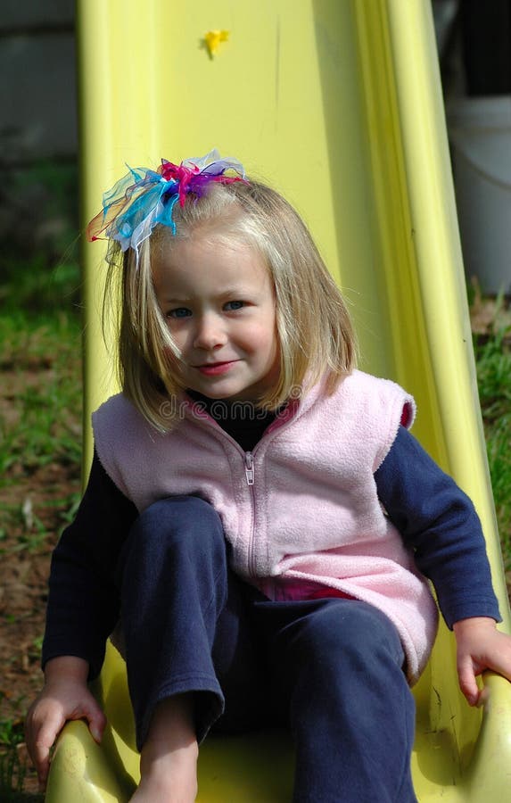 A beautiful blond caucasian white girl child with blue eyes and happy expression in her pretty face having great fun by sitting on a yellow slide and watching other kids on the playground outdoors in springtime. A beautiful blond caucasian white girl child with blue eyes and happy expression in her pretty face having great fun by sitting on a yellow slide and watching other kids on the playground outdoors in springtime