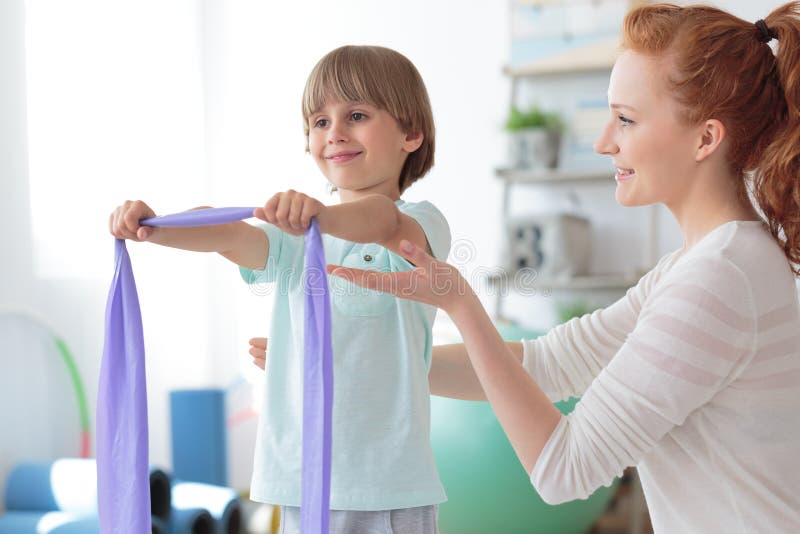 Professional physiotherapist supporting child exercising with resistance band in rehabilitation center. Professional physiotherapist supporting child exercising with resistance band in rehabilitation center