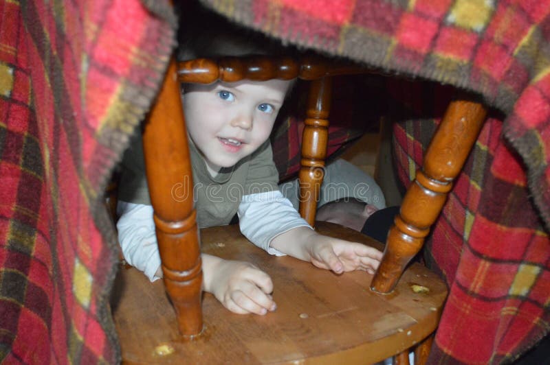 A child peeks through the chair legs that hold up his blanket fort hideaway. A child peeks through the chair legs that hold up his blanket fort hideaway.